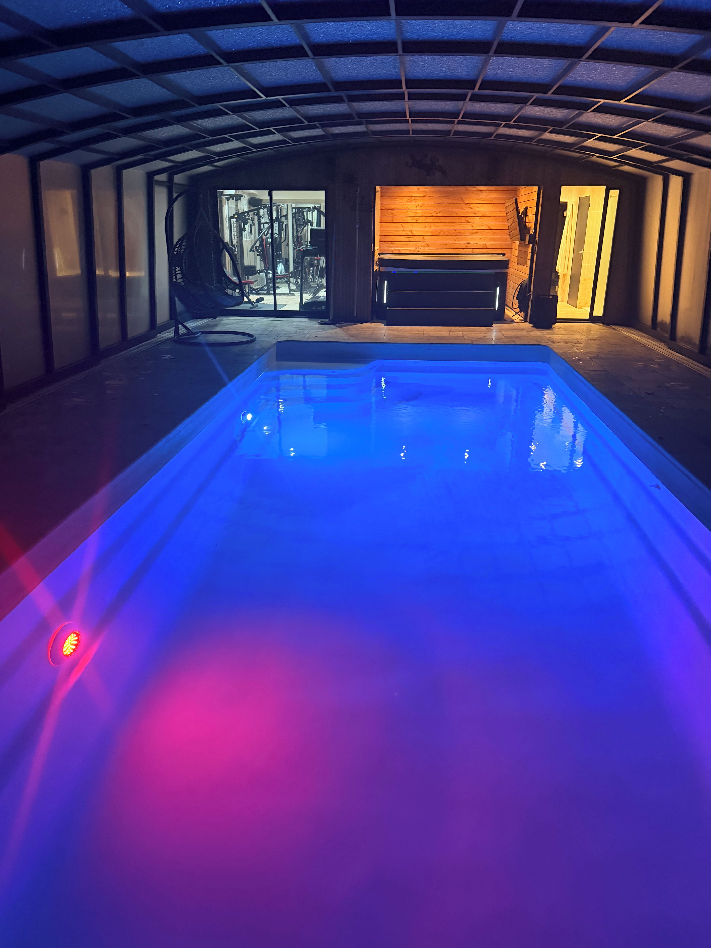 Una piscina iluminada con luz azul, un gimnasio visible a través de un ventanal y un banco de madera.