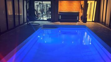 Una piscina iluminada con luz azul, un gimnasio visible a través de un ventanal y un banco de madera.