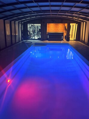 A swimming pool with a blue light, a gym visible through a glass door, and a wooden bench.
