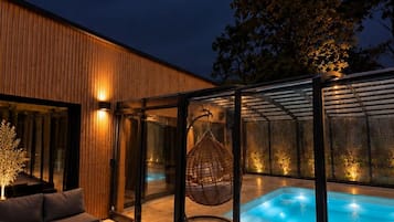 Un espace piscine extérieur moderne, doté d’une terrasse en bois, d’une structure vitrée et d’un fauteuil suspendu.