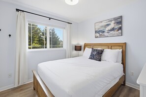 A bedroom with a bed, a window with curtains, a lamp, and a painting on the wall. - The Capella-modern, sunny, spacious, steps to beach (Pacific City)
