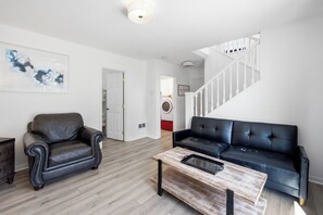 A living room with a black leather sofa, a wooden coffee table, and a black leather armchair. A staircase with white railings leads to another room. - The Capella-modern, sunny, spacious, steps to beach (Pacific City)