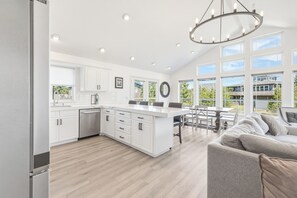 A modern kitchen with white cabinetry, a central island, and a dining area with a table and chairs. - The Capella-modern, sunny, spacious, steps to beach (Pacific City)