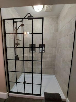 Eine Duschkabine mit Glas, eine Fliesenwand, ein Handbrausekopf und ein am Wand montierter Wasserhahn.