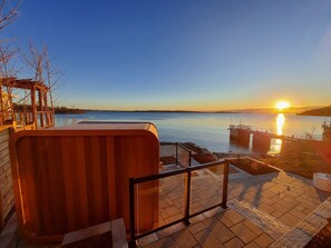 A waterfront property with a hot tub, a wooden deck, and a view of the sunset over the water.