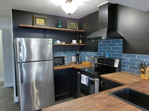Una cocina moderna con un refrigerador de acero inoxidable, un microondas, una estufa y un lavamanos. Hay estantes de madera sobre la encimera y un salpicadero de baldosas negras.