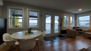 Una sala de estar moderna con una mesa redonda y sillas, un sofá, y ventanas amplias que ofrecen vista al agua.