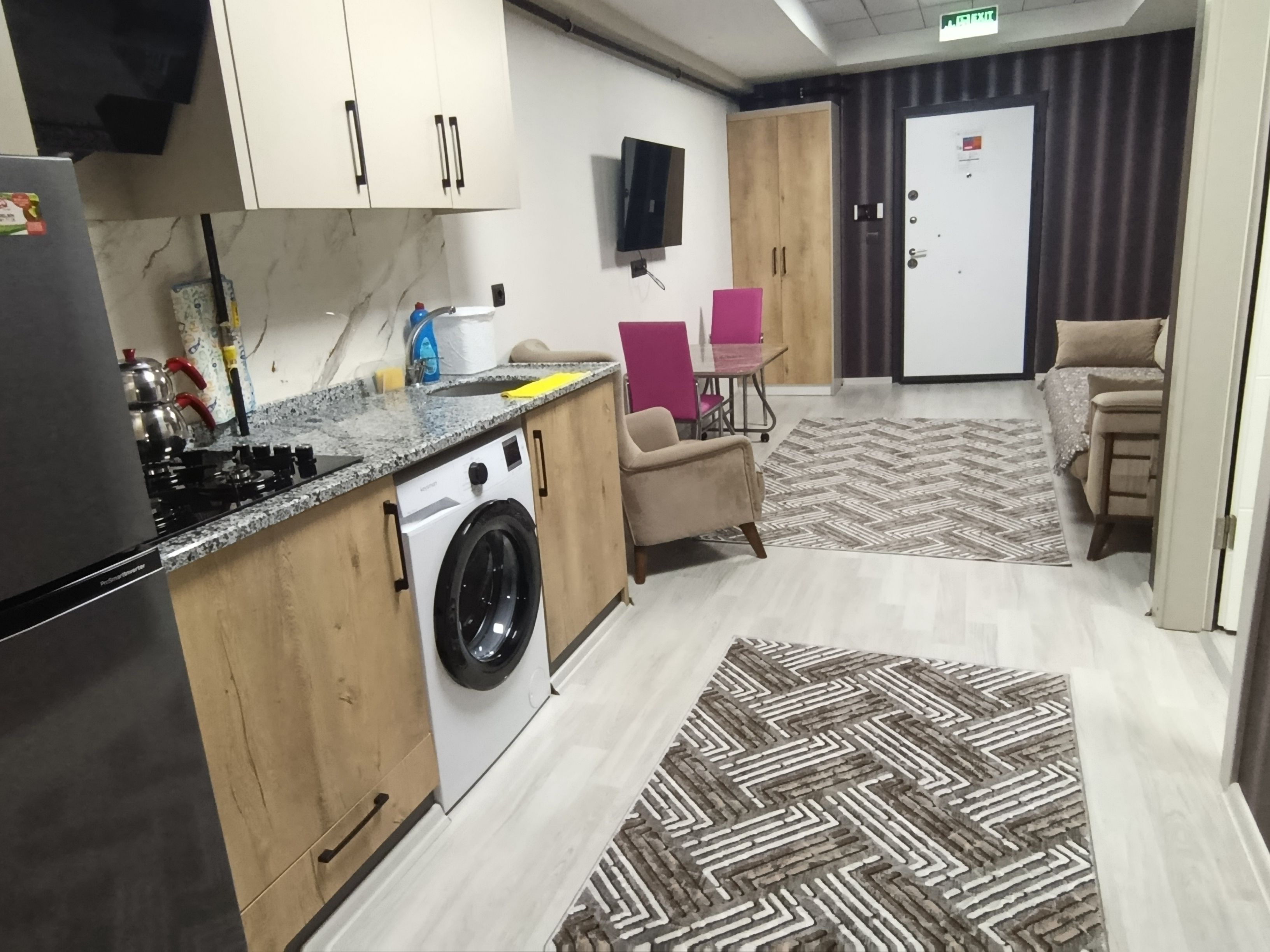 A compact kitchen with a washing machine, a stove, and a sink. There is a patterned rug on the floor and a door with a sign.