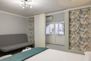 A bedroom with a grey sofa, a bed with a green blanket, and a window with a view of the outside. - Room 'Soleil Rooms - Salvia' (Catania)