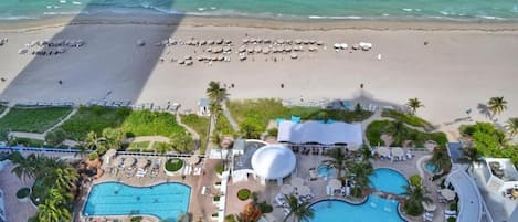 An aerial view of a resort with multiple swimming pools, a beach area with umbrellas, and a large dome structure.