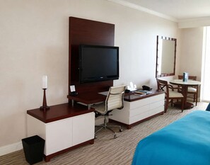 Una habitación de hotel con cama, escritorio, silla, televisor, espejo y cesto de basura.