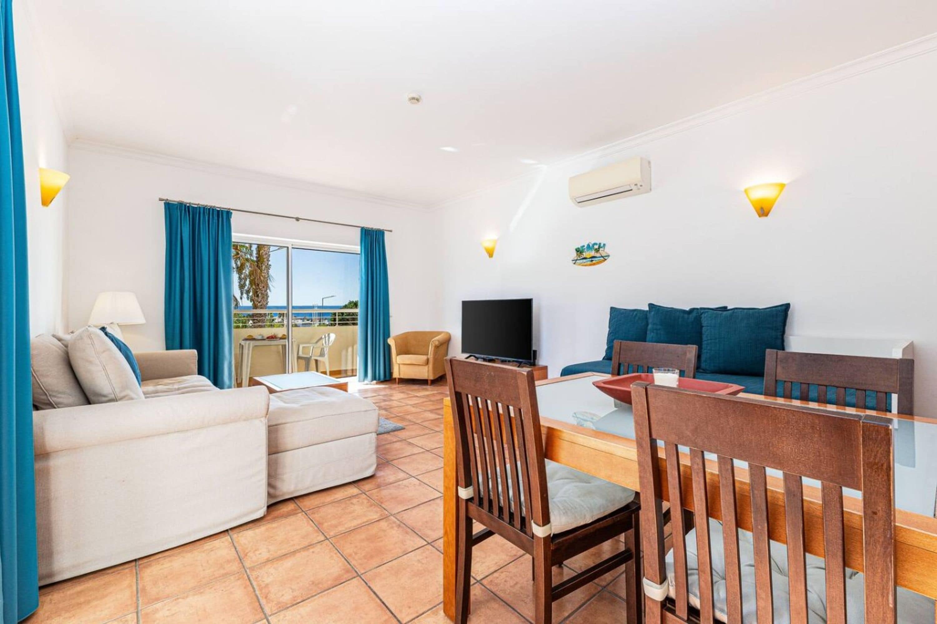 Una sala de estar con sofá, televisor, mesa de comedor con sillas y vista al mar a través de ventanas amplias.