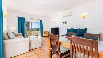 Una sala de estar con sofá, televisor, mesa de comedor con sillas y vista al mar a través de ventanas amplias.