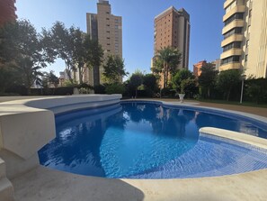 Ein Swimmingpool mit Fliesenrand, umgeben von Grünflächen und hohen Gebäuden im Hintergrund.