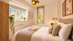 Un dormitorio con una cama, una lámpara de noche, una ventana con persianas, un cuadro en la pared y una planta.