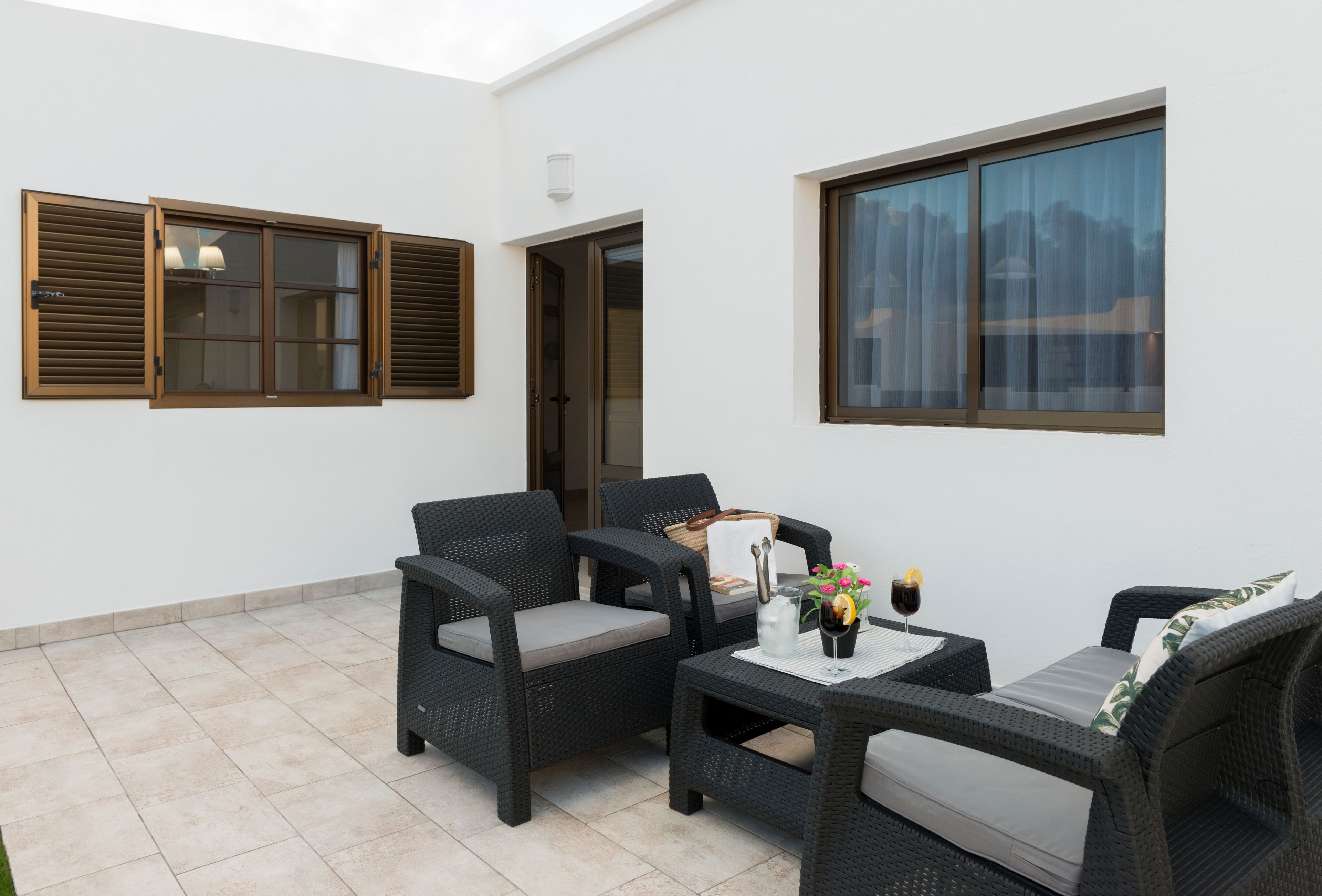 Un patio amueblado con mobiliario de mimbre, una mesa con bebidas y vistas a través de un ventanal.