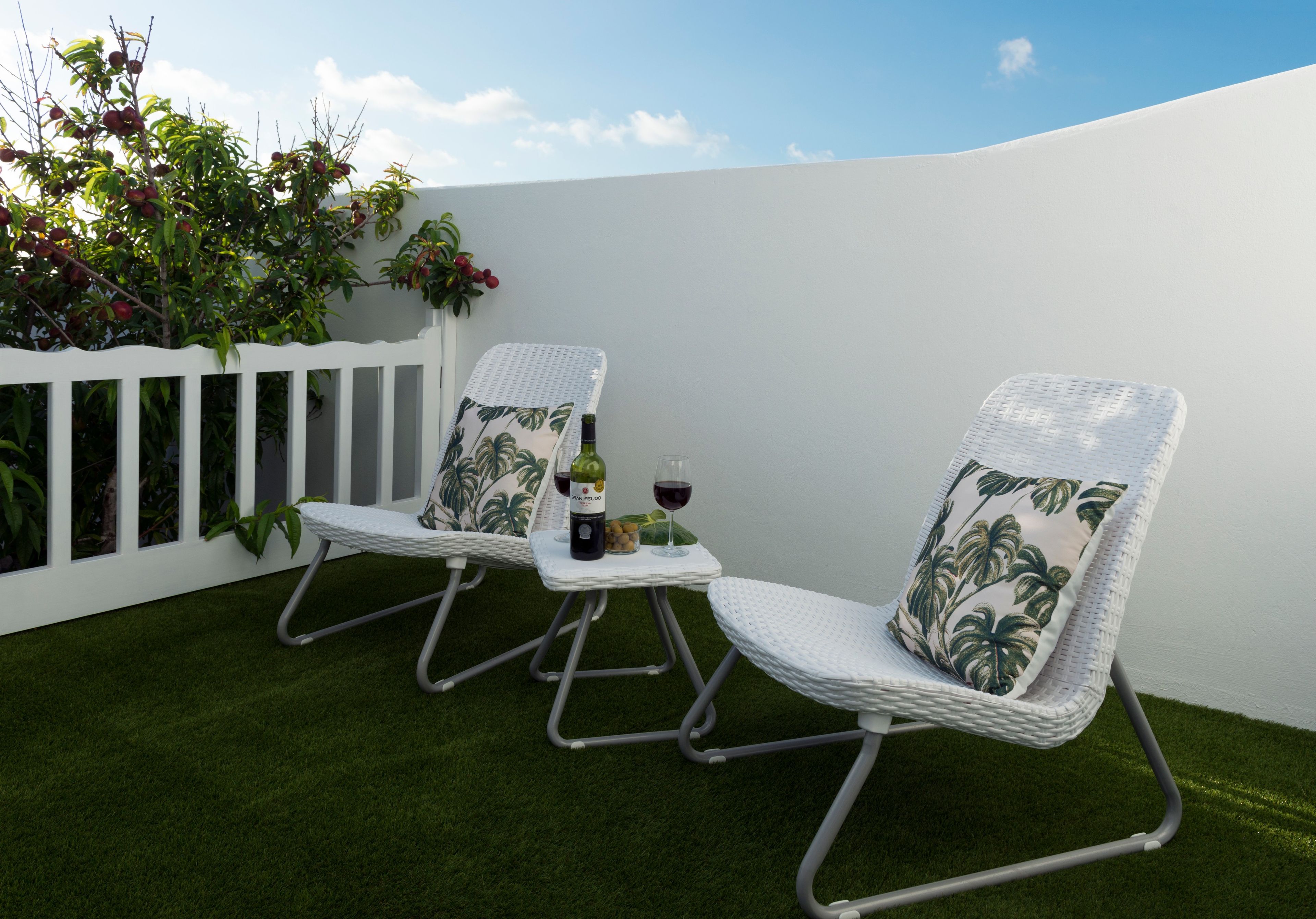 Een terras met twee witte wicker stoelen, een bijzettafeltje met een fles en glazen, en een witte schutting met beplanting.