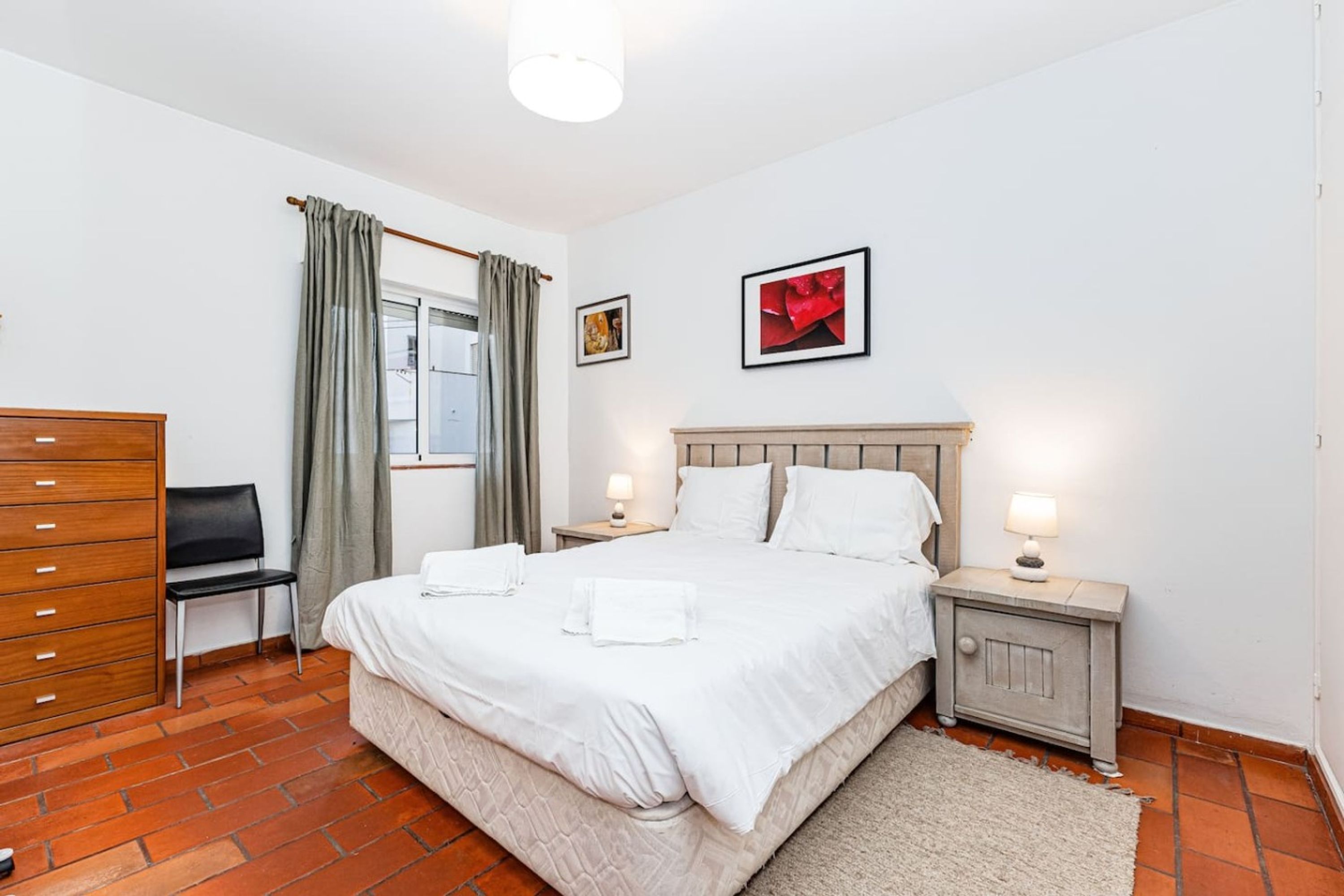 A hotel room with a bed, two bedside tables, a chair, a dresser, and a window with curtains.