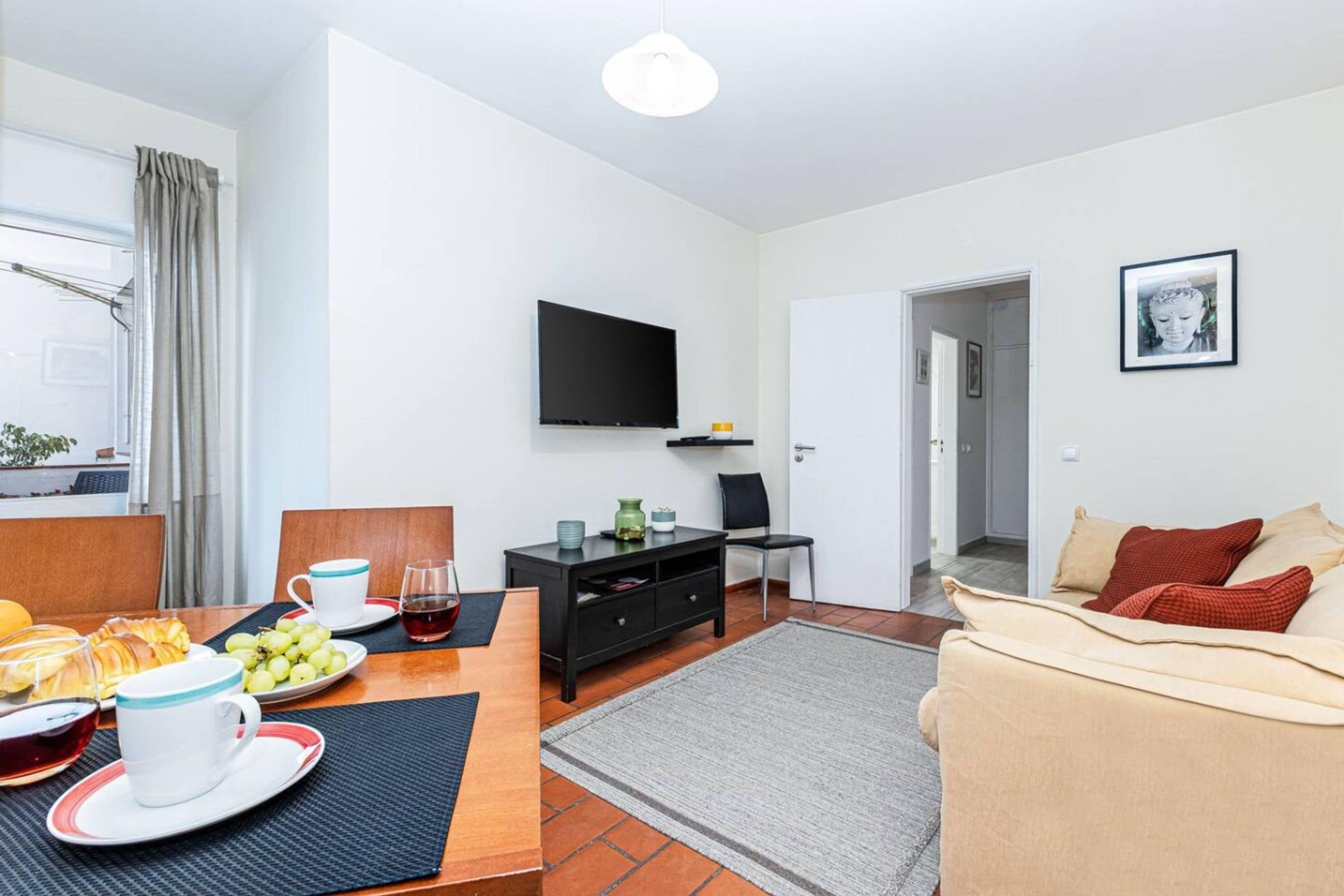A modern living room with a flat-screen TV, a dining area with a table set for two, and a sofa with cushions.