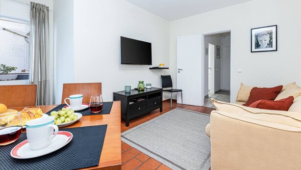 Ein modernes Wohnzimmer mit Fernseher, einem Essbereich mit Tisch für zwei Personen und einer Couch mit Kissen.