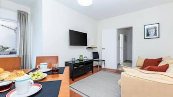 A modern living room with a flat-screen TV, a dining area with a table set for two, and a sofa with cushions.
