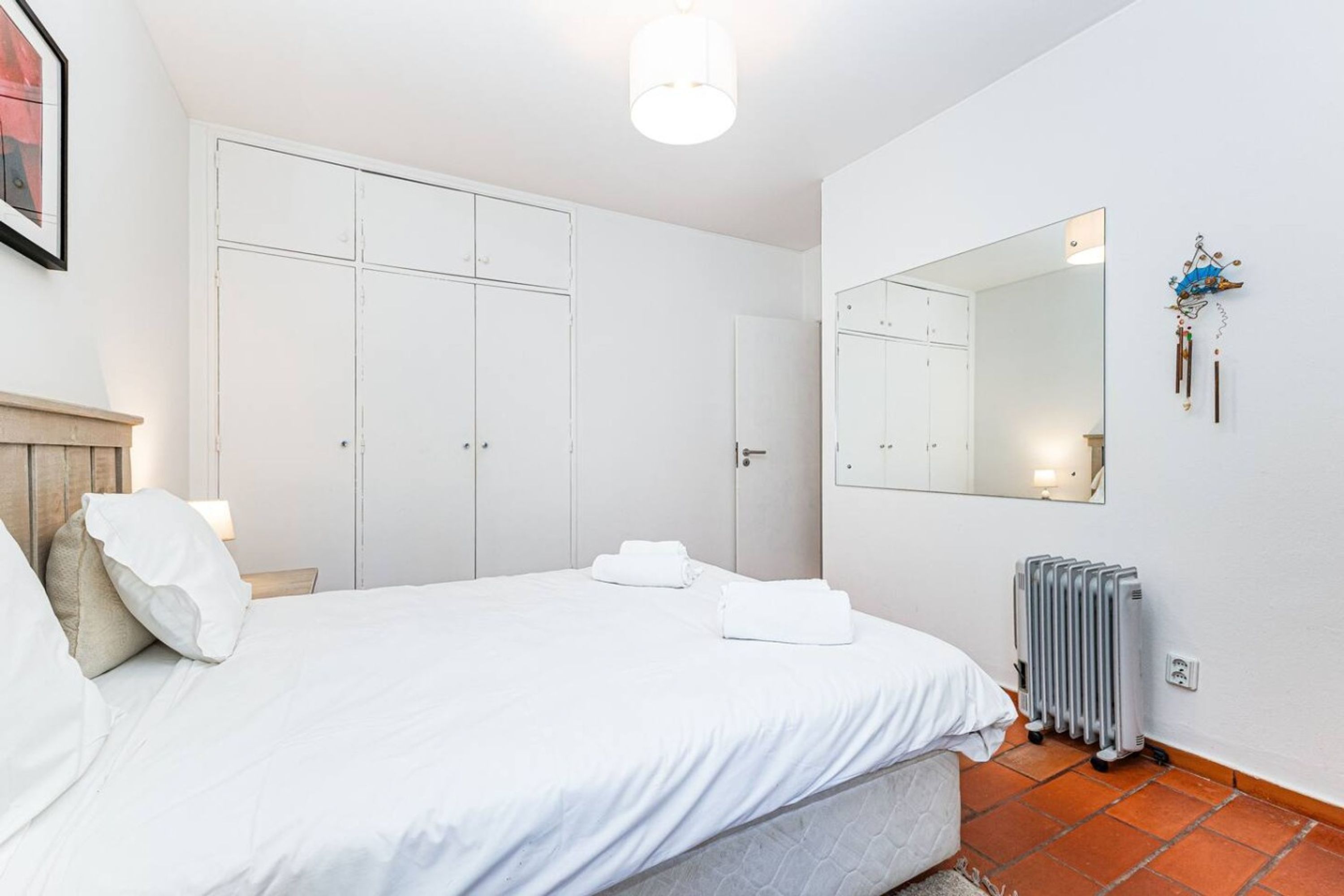 A bedroom with a bed, white curtains, a radiator, and a mirror on the wall.