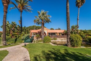A house with a paved driveway, surrounded by palm trees and a small pond. - Jardins da Meia Praia 3BR Villa Pool (Lagos)