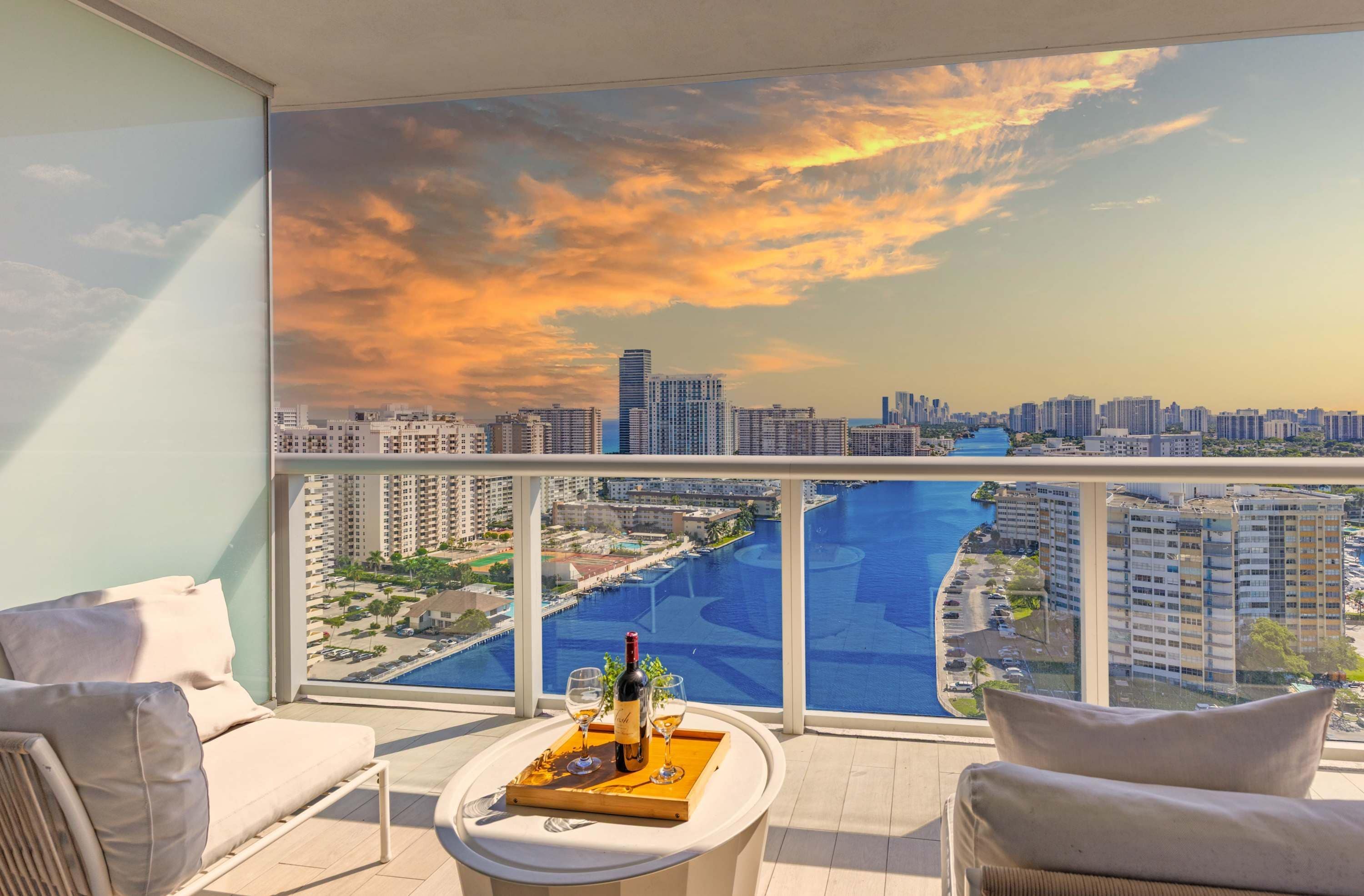 A balcony with a city view, a table with a bottle and glasses, and two chairs.