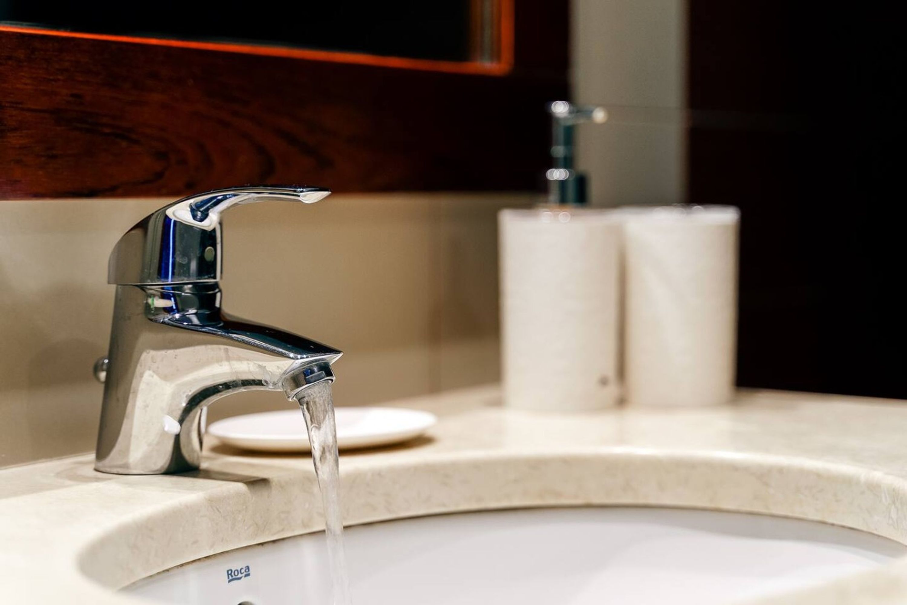 Un lavamanos con grifo de cromo y agua corriente, un dispensador de papel higiénico blanco y un espejo sobre el lavamanos.