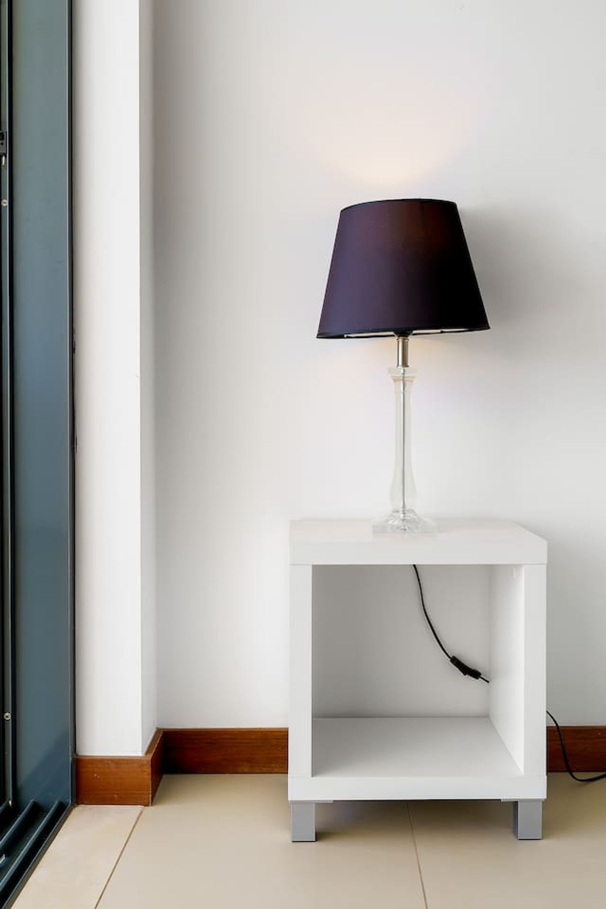 A white bedside table with a clear glass base and a black lampshade.