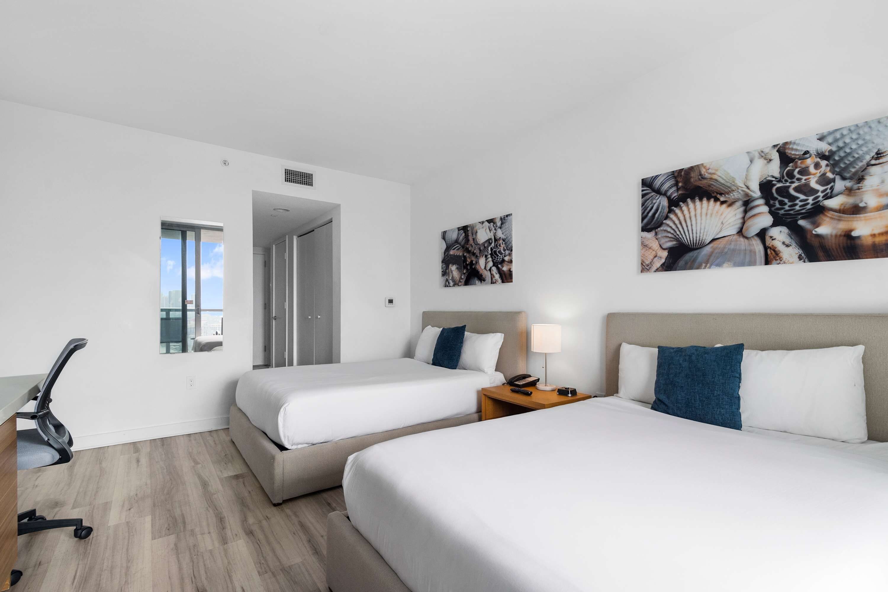 A hotel room with two beds, a desk, a chair, a lamp, and artwork on the walls.