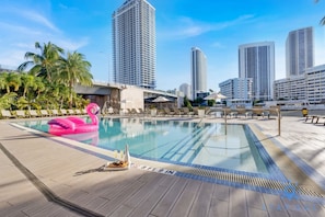 Ein Swimmingpool mit Liegestühlen und einem Flamingo-Schwimmpfaugel, umgeben von Palmen und Hochhäusern.