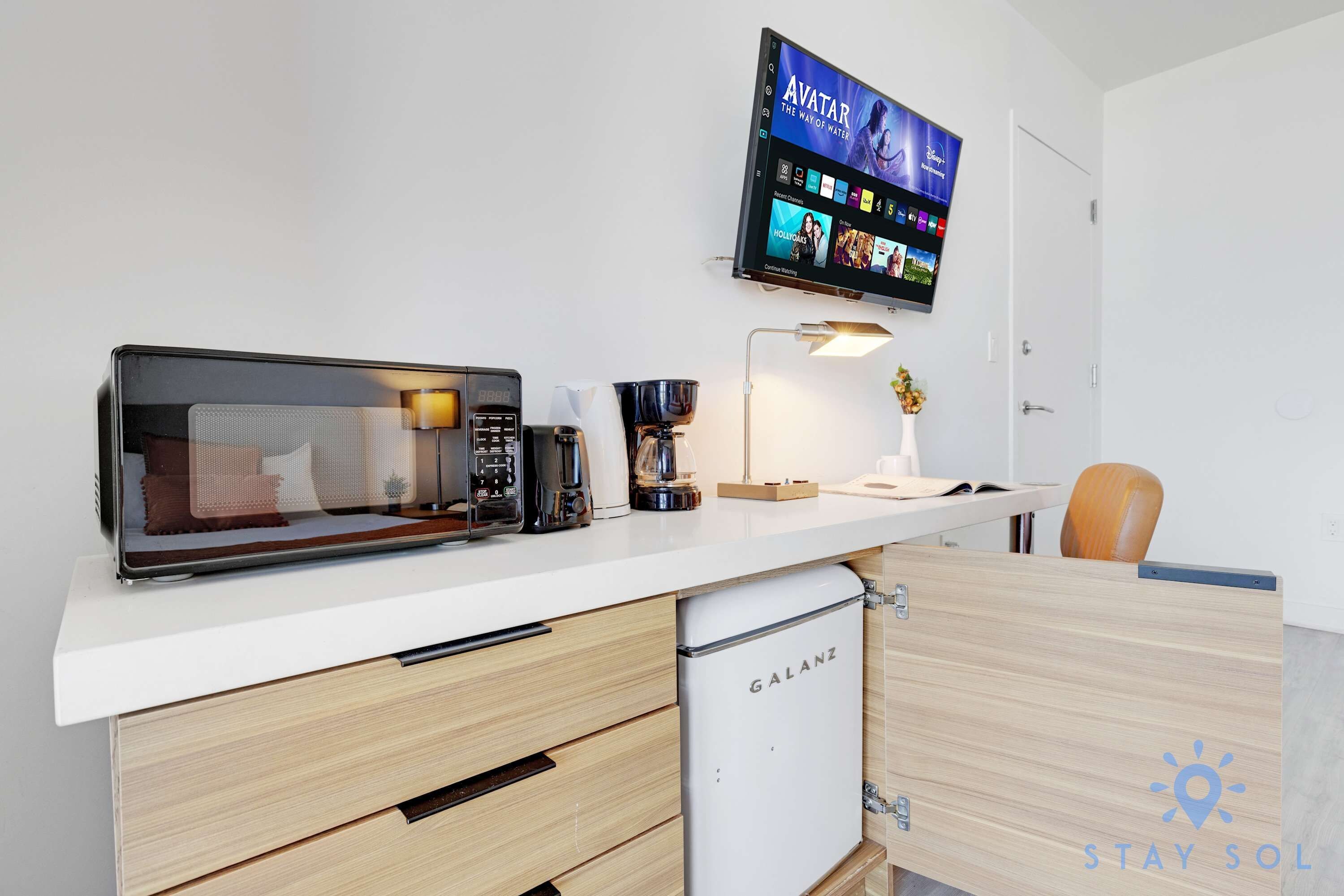 A kitchenette with a microwave, coffee maker, and a small refrigerator. A flat-screen TV mounted on the wall displays a Disney+ interface.