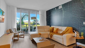 Ein modernes Wohnzimmer mit einer Couch, Sesseln und einem Couchtisch. Ein großes Fenster bietet Blick auf Palmen und Gebäude.