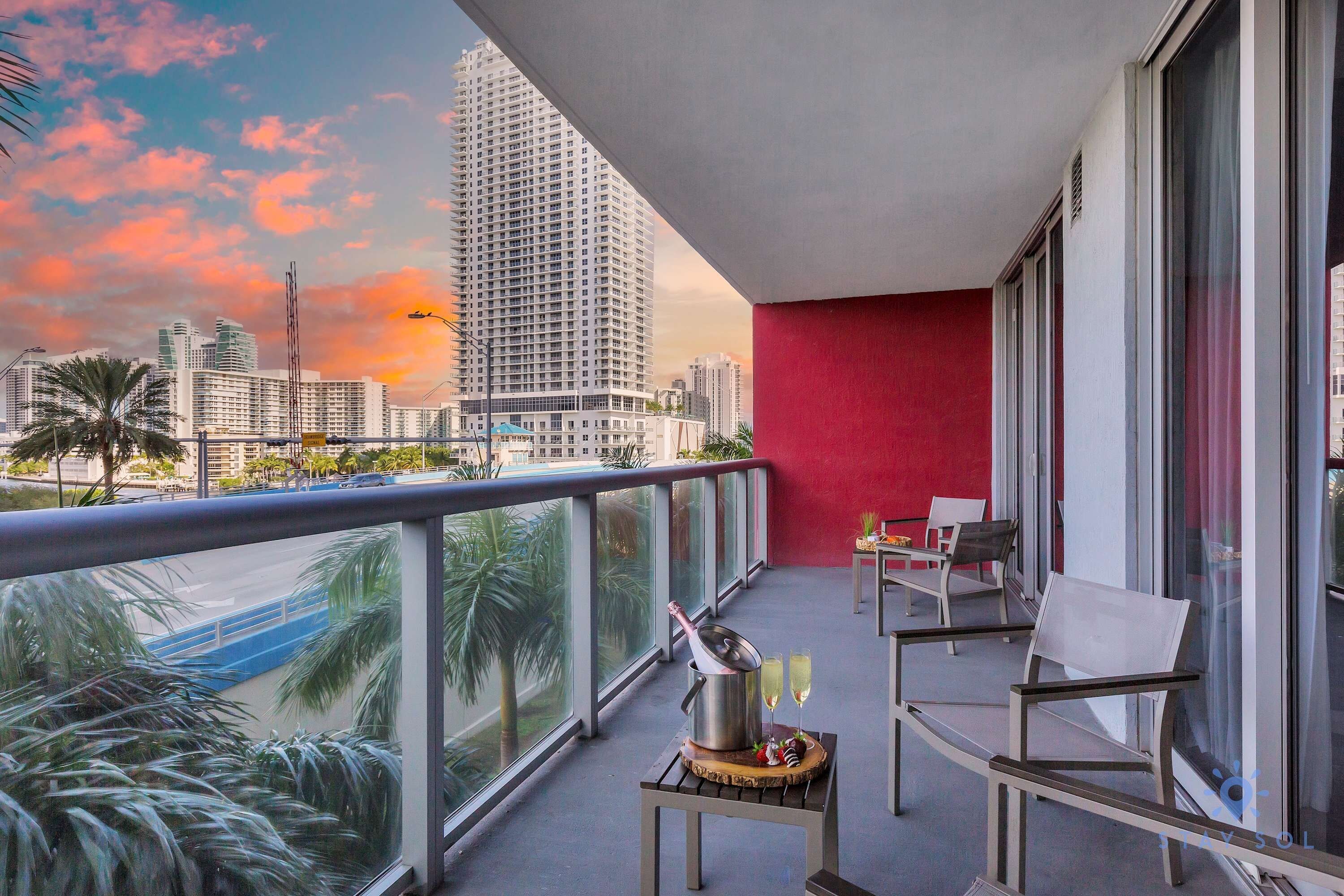 Un balcón con pared roja, una mesa con sillas y vista panorámica de la ciudad al atardecer.