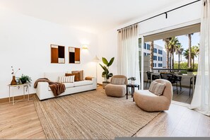 A modern living room with a sofa, armchair, and a large window with a view of palm trees and a building. - Marina Garden View Pool Marina Beach (Lagos)