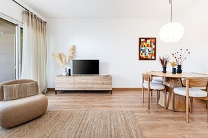 A modern living room with a flat-screen TV on a wooden cabinet, a round dining table with chairs, a woven rug, and a large window with curtains. - Marina Garden View Pool Marina Beach (Lagos)
