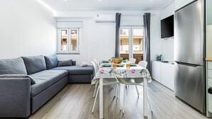 Una sala de estar moderna con un sofá seccional gris, una mesa de comedor con sillas, una zona de cocina con refrigerador y un televisor de pantalla plana montado en la pared.