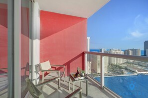 Un balcón con pared roja, una silla, una mesa con una botella y un vaso, y vistas a una ciudad y el océano.