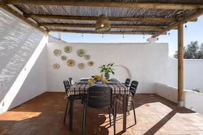 Um alpendre coberto com uma mesa posta para dois, rodeado de cadeiras e um vaso de planta na mesa.