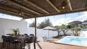 Une terrasse extérieure avec une table à manger pour quatre personnes, entourée de chaises, d’une piscine et d’un espace abrité surmonté d’une pergola.