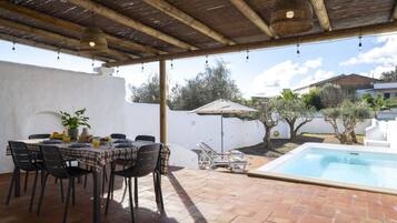 An outdoor patio with a dining table set for four, surrounded by chairs, a pool, and a covered area with a pergola overhead.