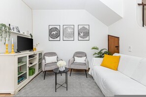 Ein modernes Wohnzimmer mit einem weißen Sofa, zwei grauen Sesseln, einem Couchtisch und einem Flachbildfernseher auf einem weißen Schrank. An der Wand hängen drei gerahmte abstrakte Kunstwerke.