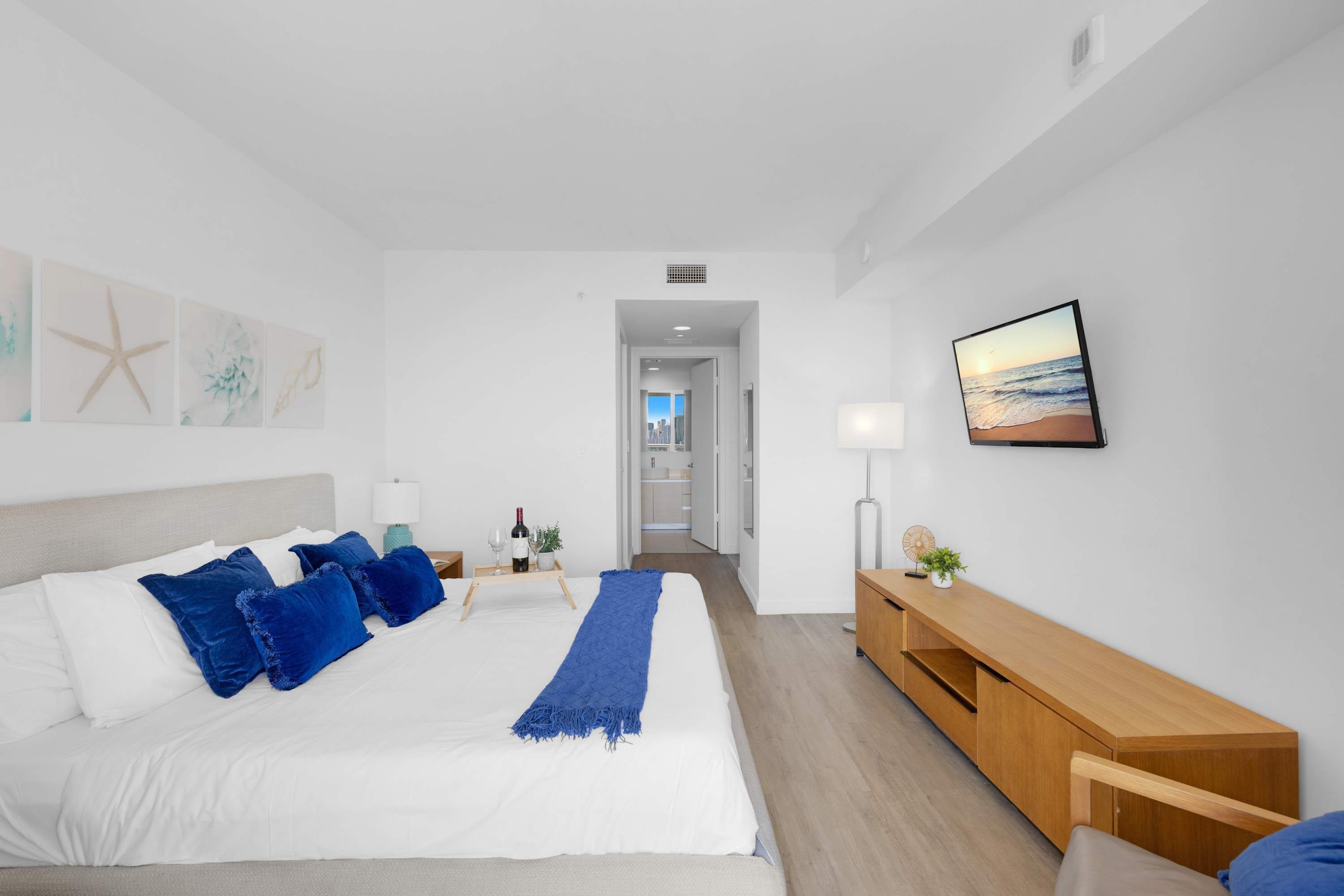 A modern hotel room with a large bed, a wooden bedside table, a TV, and a view of the city through the window.