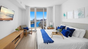 A modern hotel room with a large bed, a desk, a chair, and a view of the city and water. - Stylish Apartment 2BD 2bth (Hallandale Beach)
