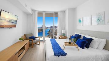 A modern hotel room with a large bed, a desk, a chair, and a view of the city and water.
