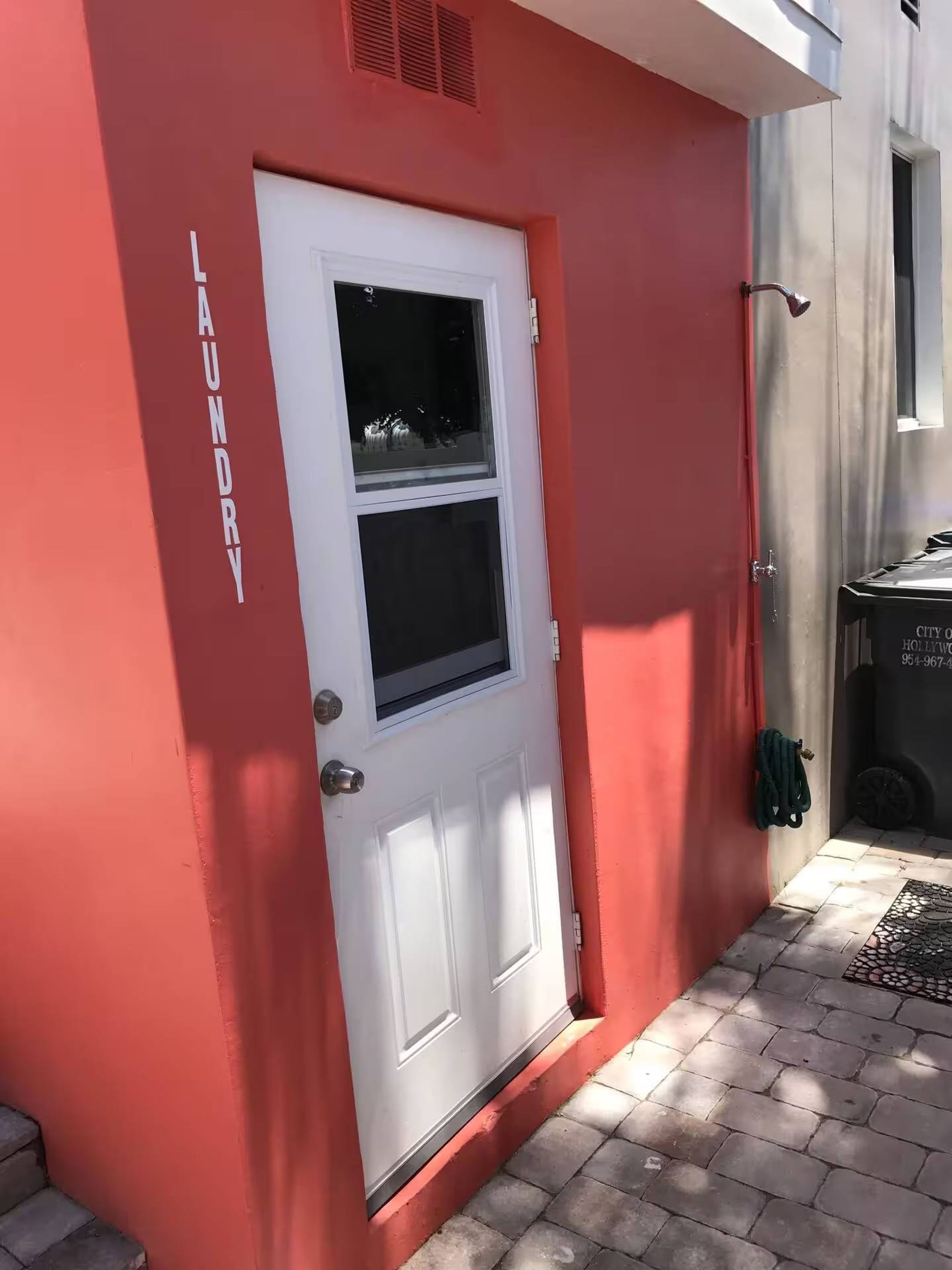 Un bâtiment rouge avec une porte blanche et une fenêtre, une poignée de porte en métal et une petite grille d’aération au-dessus de la porte.