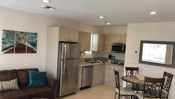 A compact living space with a kitchenette, a dining area with a round table and chairs, a sofa with cushions, and a wall-mounted TV.