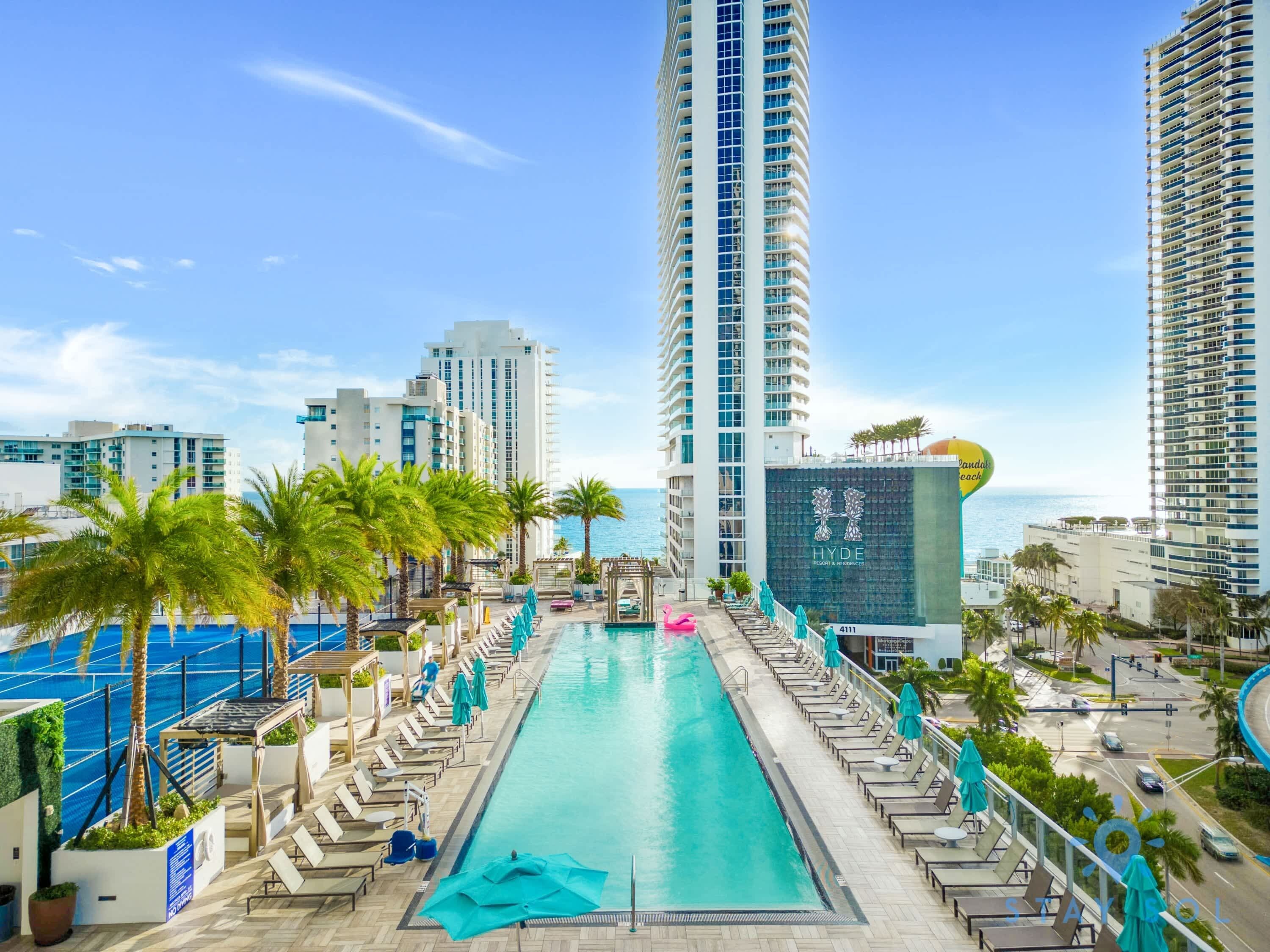 A hotel pool area with a long, rectangular pool, lounge chairs, and palm trees.