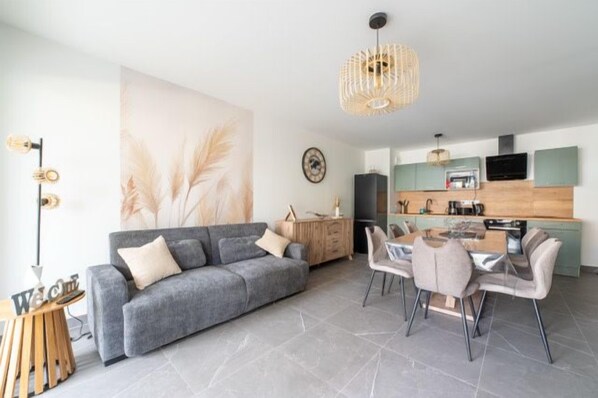 Ein modernes Wohnzimmer mit grauem Sofa, Esstisch und Stühlen, einer Küchenzeile mit grünen Schränken und einem großen Wandbild.